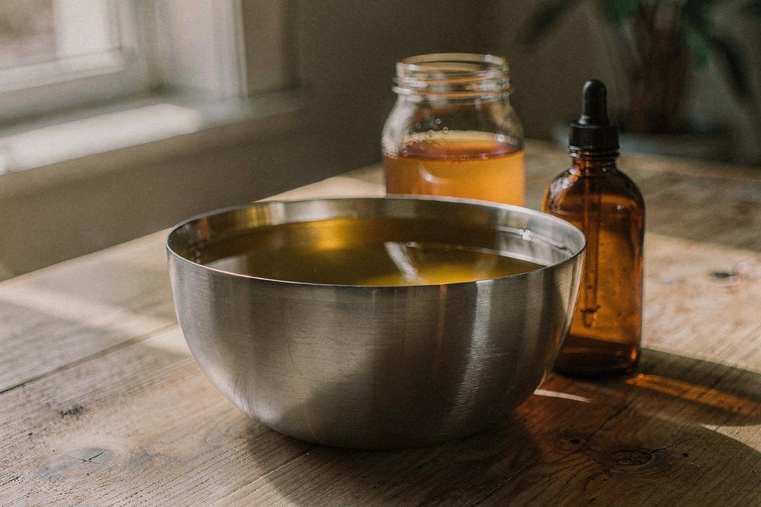 A large stainless steel bowl of golden tallow oil beside a jar of raw honey and a dropper bottle of oil