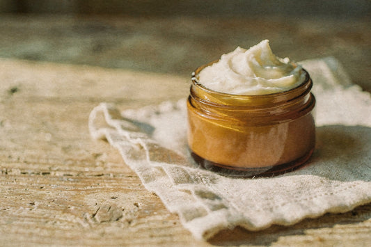 Open amber glass jar of whipped beef tallow body butter on a linen napkin on a weathered wood table