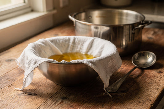 Golden tallow oil straining through cheesecloth into a stainless steel bowl, with a stockpot and ladle behind