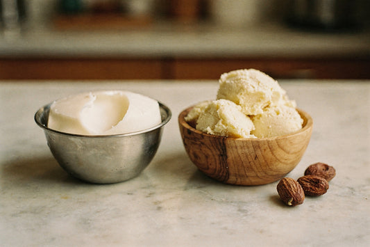 A stainless steel bowl of creamy white beef tallow beside an olive-wood bowl of raw shea butter and three dried shea nuts on a pale marble counter