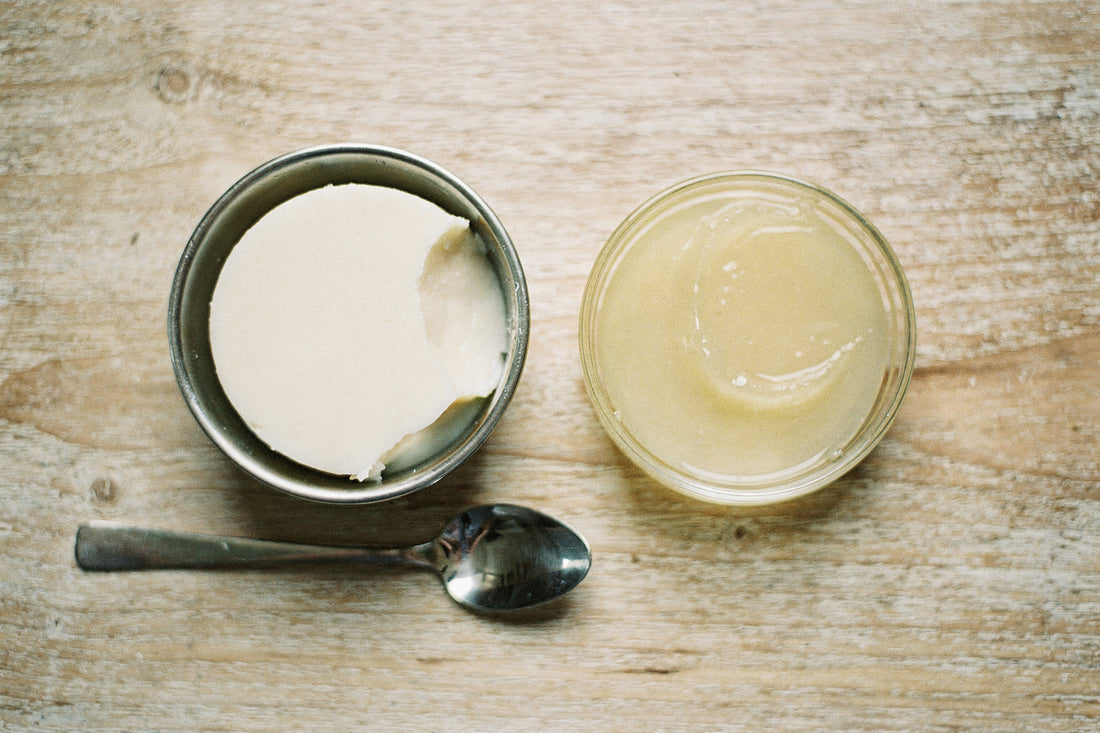 Top-down view of a steel bowl of creamy white beef tallow beside a glass dish of translucent petroleum jelly, with a spoon, on a weathered wood table