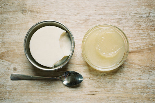 Top-down view of a steel bowl of creamy white beef tallow beside a glass dish of translucent petroleum jelly, with a spoon, on a weathered wood table