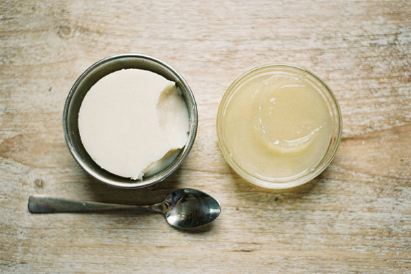 Top-down view of a steel bowl of creamy white beef tallow beside a glass dish of translucent petroleum jelly, with a spoon, on a weathered wood table