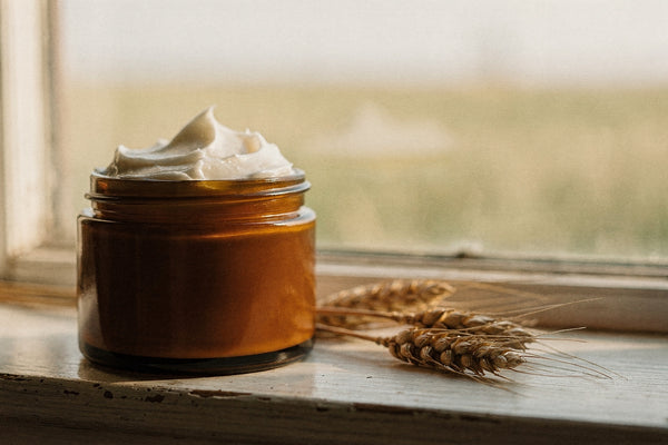 An open amber glass jar of whipped tallow body butter on a weathered window sill beside a stalk of dried wheat, with a soft field view behind